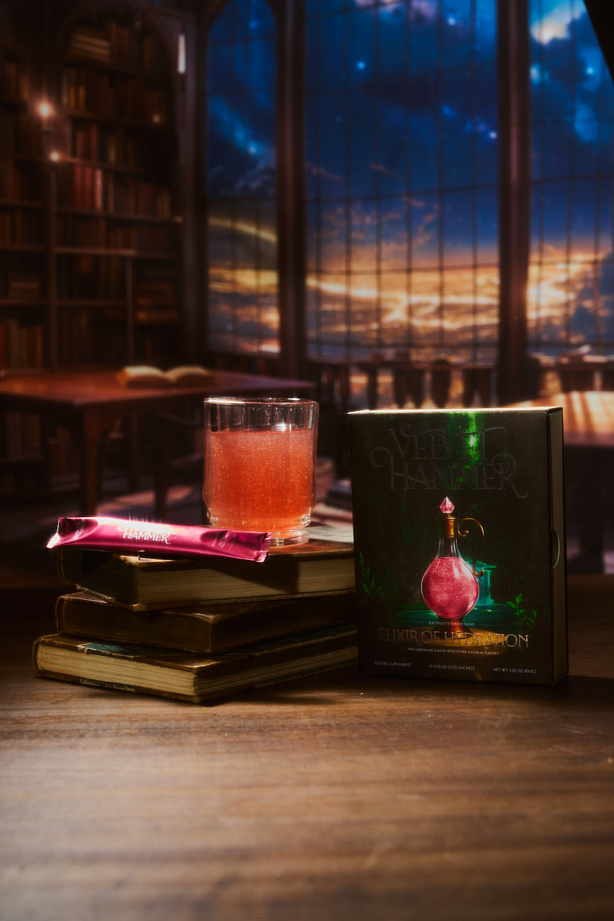 A glass of shimmering pink Elixir of Hydration on a stack of old books beside its box, in a library at dusk